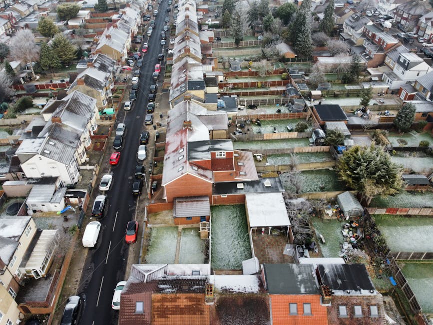 An aerial view of a residential neighborhood during winter, showcasing a street lined with terraced houses that have snow-covered roofs. The houses are primarily brick with pitched roofs, some with dormer windows, and are closely spaced. On either side of the street, there are backyards enclosed by wooden fences, featuring a variety of garden layouts with lawn areas, paved patio sections, flower beds, and mature trees. Some gardens display neatly trimmed hedges, shed structures, or garden furniture. The lawns are covered with a light dusting of snow, and pathways through the gardens are visible, some cleared of snow. In the foreground, neighboring backyards reveal different levels of garden maintenance, with tools, garden furniture, and decorative elements scattered about. The surrounding environment includes a variety of deciduous and evergreen trees, contributing texture and greenery despite the winter scene. The scene is illuminated by natural daylight, highlighting the textures of the snow, the brick surfaces, and the garden vegetation. For garden maintenance, landscaping work, or hedge trimming services, [COMPANY_NAME] can assist with maintaining the tidy appearance and health of your outdoor space.