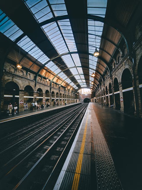 The image depicts an indoor train station with a high, arched ceiling made of glass panels supported by a black metal framework, allowing natural light to illuminate the platform below. On the left side, there are historic brick walls with multiple archways, some of which house shops and seating areas, where a few people are seen waiting or walking. The platform features a textured yellow safety line along its edge and a tactile strip for visually impaired travelers. The dark railway tracks run centrally into the tunnel at the far end, with additional tracks visible in the foreground. The environment appears clean and well-maintained, with a subdued lighting scheme complemented by mounted lamps along the brick walls. The overall scene suggests a busy yet orderly railway station, where routine maintenance and landscaping are not visible, but the structural details and atmosphere highlight a blend of functional design and historic architecture. This image does not directly relate to garden maintenance or landscaping activities but provides a detailed view of an urban transport environment. For garden-related content, reference to Gardeners Gants Hill’s outdoor services can be noted on their website at https://gardenersgantshill.org.uk.