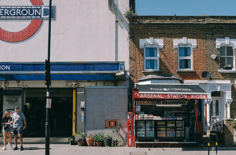 A street scene in Gants Hill showing a small kiosk labeled 'Arsenal Station Kiosk' with an open front displaying beverages and snacks. To the left, there is a partially visible underground station entrance with a large roundel sign, and two pedestrians, a man and a woman, walking past the kiosk. The surrounding buildings include a mix of brick and plaster facades, with three small white-framed windows on the brick building directly above the kiosk. Several potted plants with leafy green foliage and flowering plants are arranged outside the station entrance, adding a touch of greenery. The sidewalk appears clean and well-maintained, with a black metal post and a street lamp nearby. Bright daylight illuminates the scene, casting shadows on the ground and highlighting the textures of the brickwork, plaster, and plants. The company's gardening services, mentioned on the webpage, emphasize garden maintenance and landscaping work in the area, though not directly depicted in this street scene.