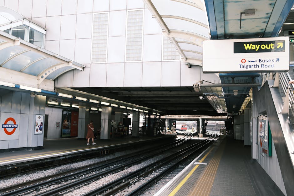 The image depicts an indoor train station with a high, arched ceiling made of glass panels supported by a black metal framework, allowing natural light to illuminate the platform below. On the left side, there are historic brick walls with multiple archways, some of which house shops and seating areas, where a few people are seen waiting or walking. The platform features a textured yellow safety line along its edge and a tactile strip for visually impaired travelers. The dark railway tracks run centrally into the tunnel at the far end, with additional tracks visible in the foreground. The environment appears clean and well-maintained, with a subdued lighting scheme complemented by mounted lamps along the brick walls. The overall scene suggests a busy yet orderly railway station, where routine maintenance and landscaping are not visible, but the structural details and atmosphere highlight a blend of functional design and historic architecture. This image does not directly relate to garden maintenance or landscaping activities but provides a detailed view of an urban transport environment. For garden-related content, reference to Gardeners Gants Hill’s outdoor services can be noted on their website at https://gardenersgantshill.org.uk.