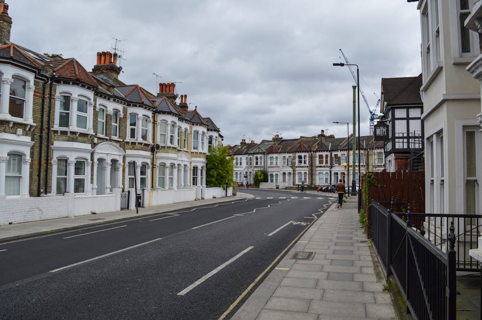 A quiet residential street along Ilford High Road with a row of Victorian-style terraced houses, featuring ornate white facades, decorative window trims, and reddish-brown tiled roofs with multiple chimneys. The pavement on the right side is made of gray paving slabs, bordered by a black wrought-iron fence and a small garden area with lush green shrubbery. The street is paved with dark asphalt marked with white lines for parking and traffic lanes. Overhead, there are streetlights and a construction crane in the distance, set against a cloudy, overcast sky. The scene captures an urban environment with well-maintained properties, typical of Gants Hill, with no visible garden or outdoor maintenance activity, but reflects the area served by Gardeners Gants Hill for landscaping and gardening services.